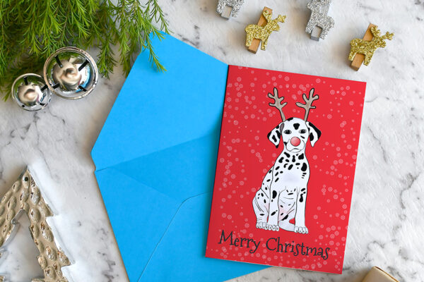 Red Christmas card featuring a cartoon Dalmatian wearing reindeer antlers, on a blue envelope, surrounded by Christmas decorations.