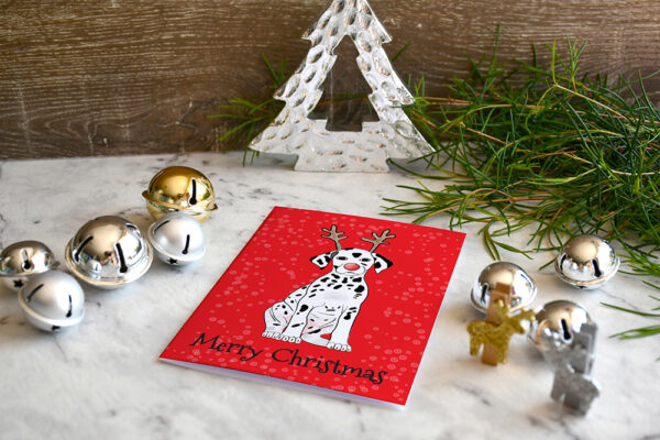 Red Christmas card featuring a cartoon Dalmatian wearing reindeer antlers, surrounded by Christmas decorations and greenery.