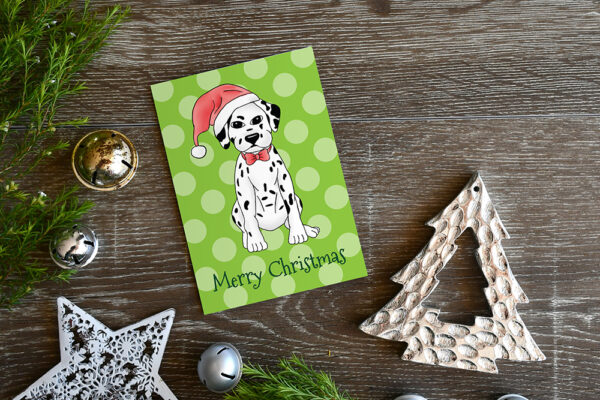 Polka dot green Christmas Card featuring a cartoon Dalmatian in a Santa hat, on a wooden surface surrounded by Christmas decorations and greenery.