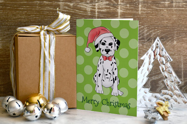 Green polka dot Christmas card featuring a cartoon Dalmatian in a Santa hat between a gift and a Christmas tree decoration with baubles scattered around.