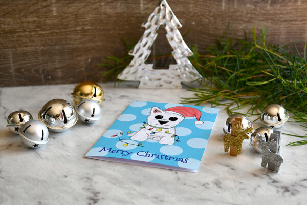 Blue polka dot Christmas card featuring Westie, Alf, in a Santa hat and wrapped in Christmas lights on a marble and wood background surrounded by Christmas decorations and greenery.