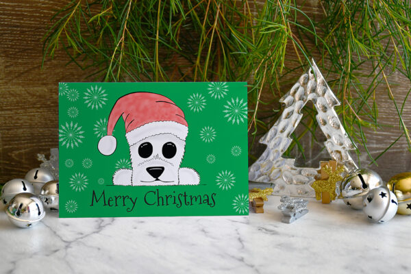 Green Christmas card featuring cartoon Westie in a Santa hat on a marble and wood background surrounded by Christmas decorations and greenery.