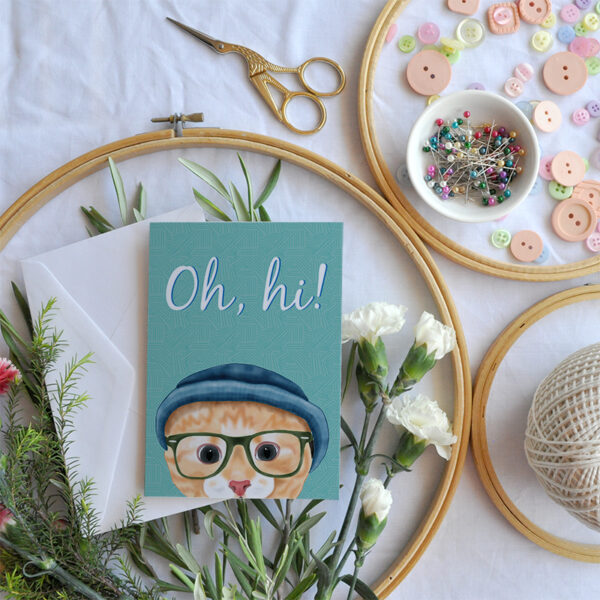 A card featuring a cartoon hipster Ginger cat in a beanie and glasses on a white background with sewing accessories and greenery around it.