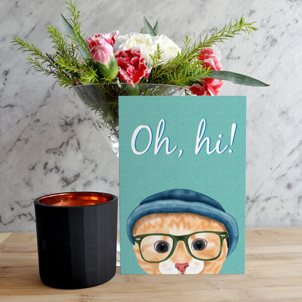 A card featuring a cartoon hipster Ginger cat in a beanie and glasses with a marble background next to a black candle with a vase of flowers behind it.