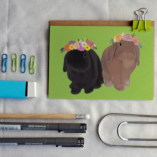 A greeting card featuring a black rabbit and brown rabbit, Indy and Dash, in flower crowns on a green background, placed amongst stationery items.