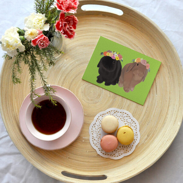 A greeting card featuring a black rabbit and brown rabbit, Indy and Dash, in flower crowns on a green background, placed on a wooden tray with a cup of tea, macaroons and vase of flowers.