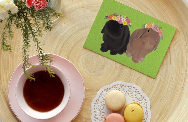 A greeting card featuring a black rabbit and brown rabbit, Indy and Dash, in flower crowns on a green background, placed on a wooden tray with a cup of tea, macaroons and vase of flowers up close.