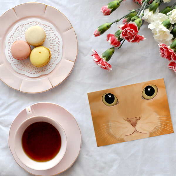 A card featuring a cartoon picture of a close up ginger cat. The card is on a white background with a cup of tea, plate of macarons and bunch of flowers.