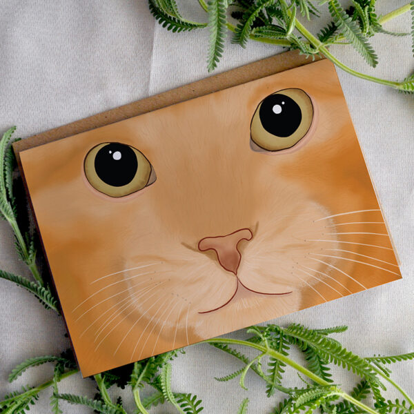 A card featuring a cartoon picture of a close up ginger cat. The card is on a white background with greenery.